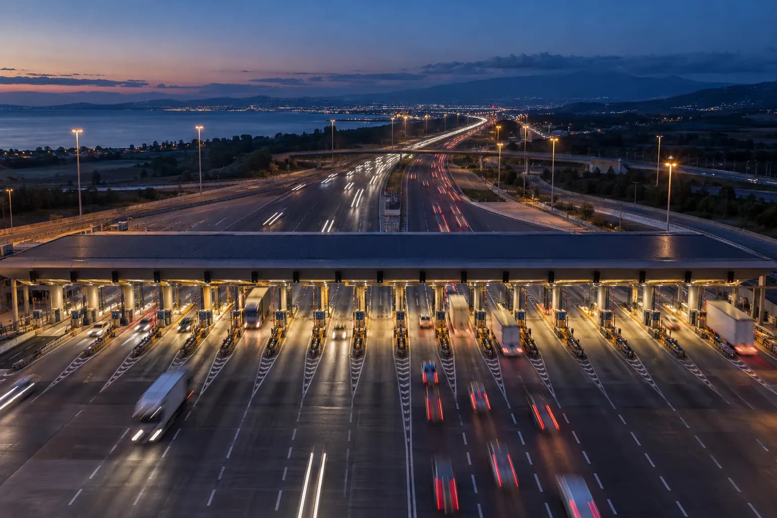 Motorway toll plaza at dusk seen from above, with barriers and vehicle light trails